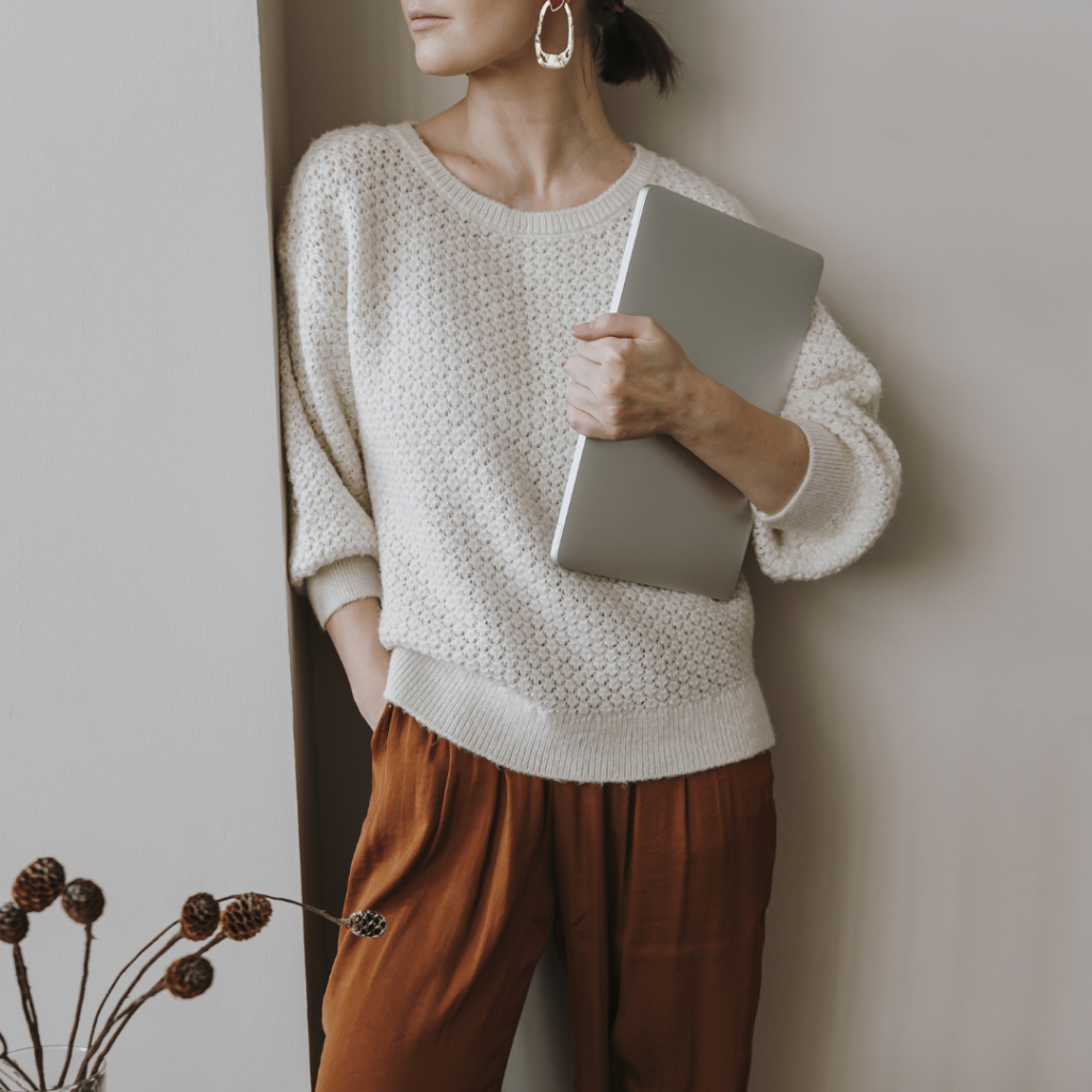 A woman in a white sweater and rust-colored pants holds a closed laptop against her side, standing beside a wall with dried flowers in the foreground.