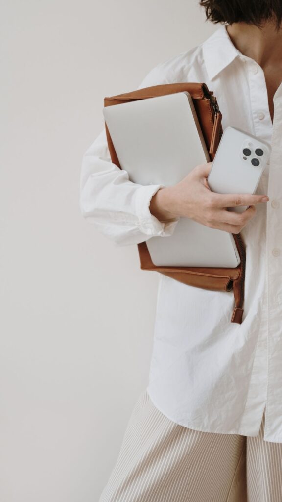 A person in a white shirt holding a brown leather bag and a smartphone, against a neutral background.