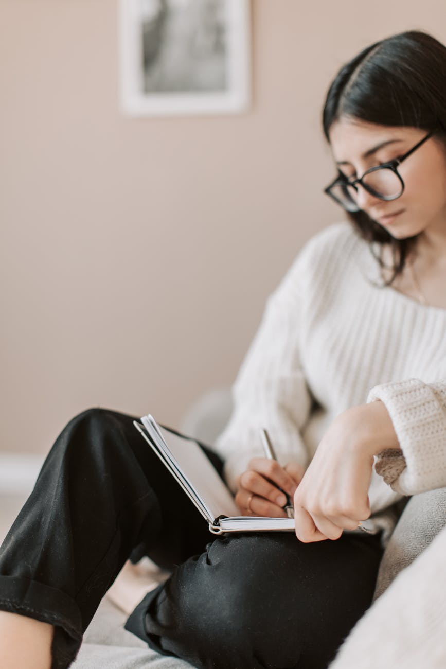 crop young woman writing schedule in diary on sofa