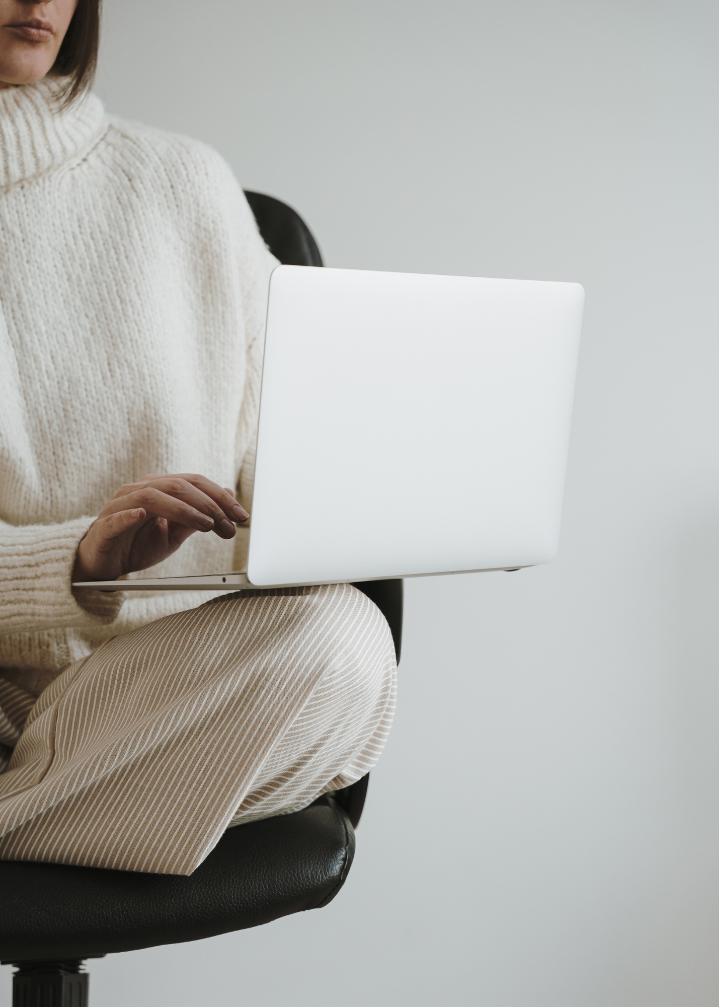 Small business owner sitting in a chair working on her laptop. She is wearing a comfy white knit sweater and white slacks